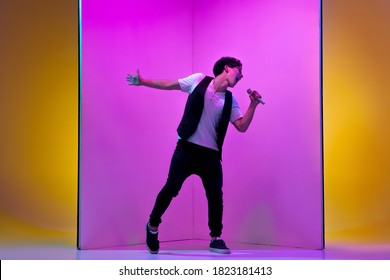 Young Male Musician, Singer Performing On Pink-orange Background In Neon Light. Concept Of Music, Hobby, Festival, Entertainment, Emotions. Joyful Party Host, Singer, Portrait Of Artist.