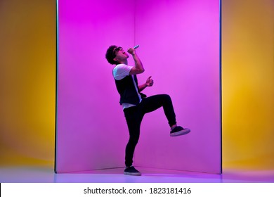 Young Male Musician, Singer Performing On Pink-orange Background In Neon Light. Concept Of Music, Hobby, Festival, Entertainment, Emotions. Joyful Party Host, Singer, Portrait Of Artist.