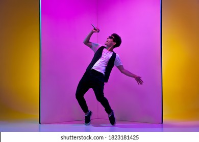 Young Male Musician, Singer Performing On Pink-orange Background In Neon Light. Concept Of Music, Hobby, Festival, Entertainment, Emotions. Joyful Party Host, Singer, Portrait Of Artist.