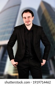 Young Male Office Manager Posing In Front Of Mirrored Skycrapers. He Looks Confident While Holding His Hips With His Hands. Man Wears Black Strict Suit. Financial District In The Background.