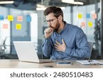 A young male office worker sits at a desk at work and coughs, covers his mouth with his hand and holds his chest, breathing hard and feeling unwell.