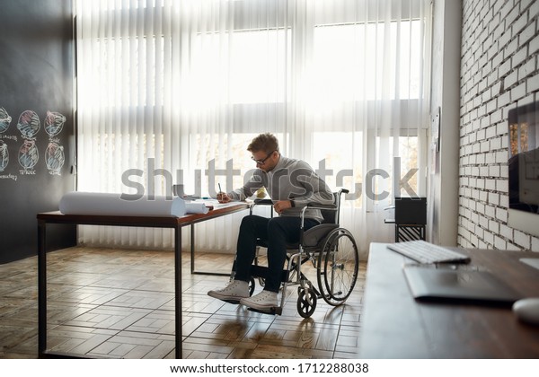 Young\
male office worker in a wheelchair drawing something while sitting\
at his workplace in the modern bright office. Disability and\
handicap concept. Construction and\
architecture