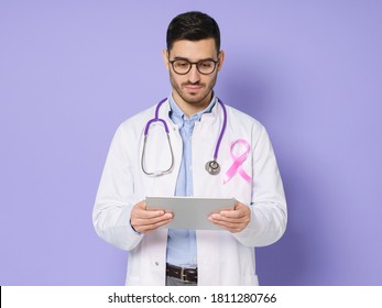 Young Male Oncologist Holding Tablet And Looking Attentively At Screen, Wearing Pink Ribbon On His Robe, Feeling Responsible, Isolated On Purple Background