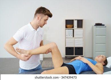 Young Male Orthopedist Massaging The Impaired Leg Of A Female Patient Who Is Lying Down On A Therapy Bed.