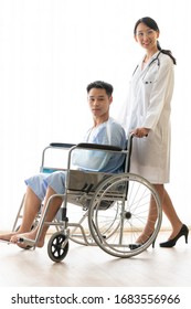 Young Male Patient Who Broken Arm From Accident Sitting On Wheelchair Looking At Camera. Full Length Body Of Beautiful Long Hair Female Doctor Smiling. Isolated White Background Copy Space.