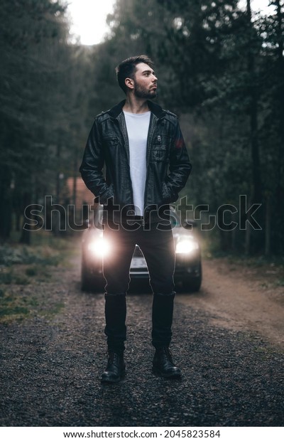 Young, male person in leather jacket standing\
in front of his car in the middle of the woods. Portrait of\
bearded, stylish man looking to the\
side.