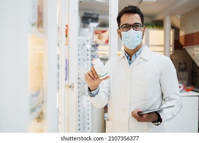 Young Male Pharmacist With Face Protective Mask Working In A Drugstore. He Is Smiling And Holding Some Drugs While Looking At Camera.