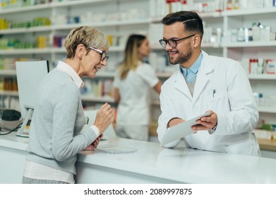 Young male pharmacist giving prescription medications to senior female customer in a pharmacy with female pharmacist in the background - Shutterstock ID 2089997875