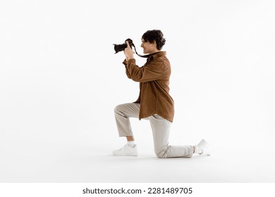 Young male photographer in classic outfit, holding his professional camera and taking photo, standing on one knee on white studio background, side view - Powered by Shutterstock