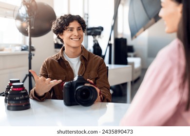 Young male photographer with curly hair, holds DSLR camera while engaging in light-hearted discussion with female model, in sunny photo studio environment - Powered by Shutterstock