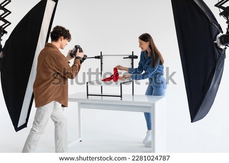 Young male photographer taking photo of stylish red shoes on white platform background in photostudio with modern equipment, lady helping to make content photoshoot