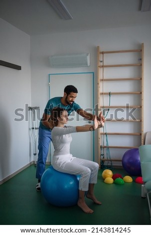 Young male physiotherapist exercising with young woman patient on ball in a physic room