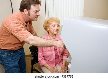 Young Male Poll Worker Assisting A Senior Woman With A New Touch Screen Voting Machine.