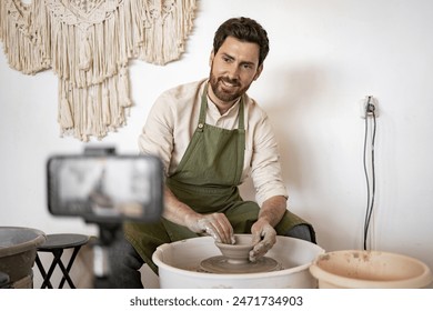 Young male potter creating ceramic vase on potter's wheel while recording video blog using smartphone. Showcasing craftsmanship and creativity in pottery. - Powered by Shutterstock