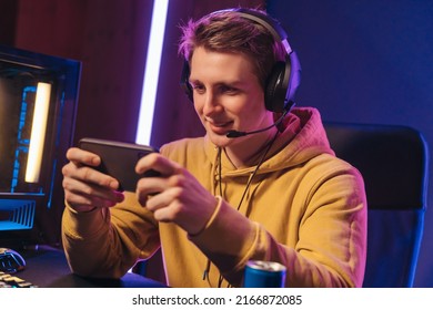 Young male professional cybersport gamer playing mobile game by smartphone on online tournament in neon lights room. Smiling pro gamer wearing headset streaming while participates in eSport event
