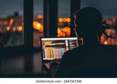A young male programmer in front of a window with a lights of night city, on his laptop screen is code. out of focus