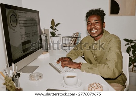 Young male programmer sitting at table and typing on keyboard while posing for portrait in office