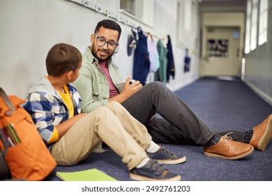 Young male psychologist speaking to depressed boy in school while sitting on the floor in school corridor. Man working with a child while discussing about learning problem and bullying. - Powered by Shutterstock