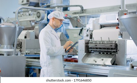 Young Male Quality Supervisor Or Food Technician Is Inspecting The Automated Production At A Dumpling Food Factory. Employee Uses A Tablet Computer For Work. He Wears White Sanitary Hat And Work Robe.