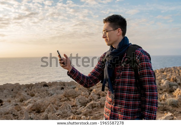 Young male in a remote location on a rocky\
cliff with sea on background. Man holding smartphone and seeking\
network connection.