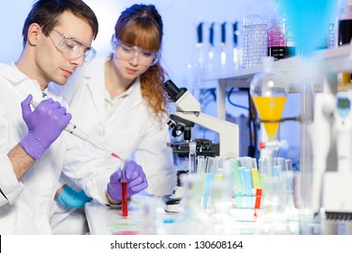 Young male researcher pipetting red liquid in the glass tube in the (forensics, microbiology, biochemistry, genetics, oncology...) laboratory. Female assistant science student learning the protocol. - Powered by Shutterstock