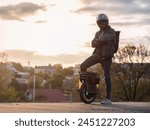 A young male rider in protective gear with a backpack on an electric unicycle against the background of the city at sunset. Driving around the city on a mono wheel