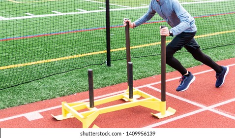 A Young Male Runner Is Pushing A Yellow Sled With No Weight On It Down A Red Track During Practice.