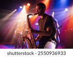 Young male saxophonist playing saxophone during live performance on stage, bandmates playing instruments in background, colorful stage lights illuminating scene