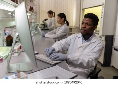 Young Male Scientist In Gloves And Whitecoat Sitting In Front Of Computer Screen In Lab