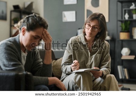 Young male sharing personal issues with female therapist during counseling session, therapist taking notes, room filled with bookshelves and decorations
