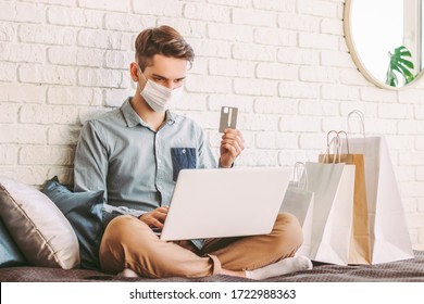 Young Male Shopper In Medical Face Mask Using Laptop On Couch For Online Shopping. Hipster Man In Protective Mask Hold Credit Card In Hand. Digital Payment. E-commerce Store. COVID-19 Home Quarantine
