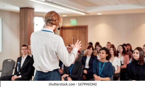 Young male speaker in suit with headset giving a talk at business meeting. Audience listening at conference hall. Selective focus. Horizontal shot