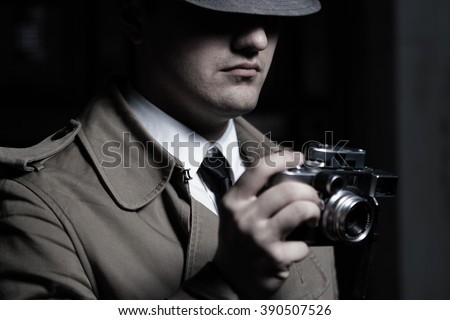 Young male spy agent wearing a hat, coat and a tie, holding rangefinder Leica film camera. Eyes hidden in the shadow.