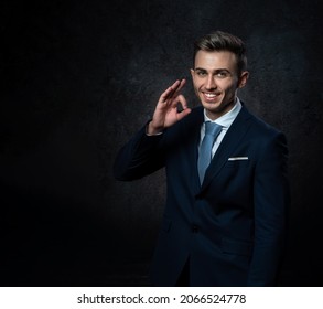 A Young Male Steward, In A Suit, Smiles, Makes A Hand Gesture Everything Is Ok. Against A Dark Background.