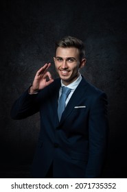 A Young Male Steward, In A Suit, Smiles, Makes A Hand Gesture Everything Is Ok. Against A Dark Background.