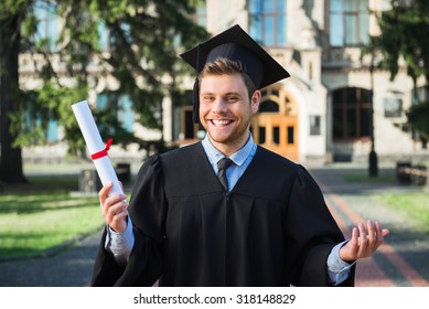 Young Male Student Dressed In Black Graduation Gown. Campus As A Background. Boy Cheerfully Smiling, Holding Diploma And Looking At Camera