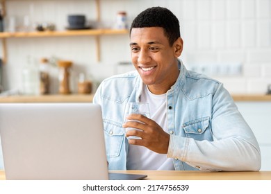 Young Male Student Or Freelancer Uses Laptop For Online Education Or Work, Drinks Fresh Water. Happy African American Man Working From Home, Studying, Distance Learning