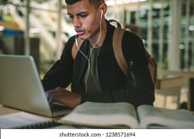 Young Male Student Preparing School Assignment In Library. Man With Earphones And Bag Working On Laptop At College Library.