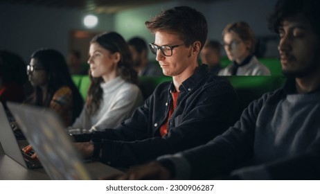 Young Male Student Studying in University with Diverse Multiethnic Classmates. He is in Uplifting Mood and is Using a Laptop Computer. Patiently Listening to Professor's Lecture and Taking Notes - Shutterstock ID 2300906287