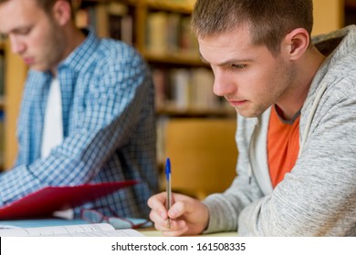 Young Male Students Writing Notes At Desk In The College Library