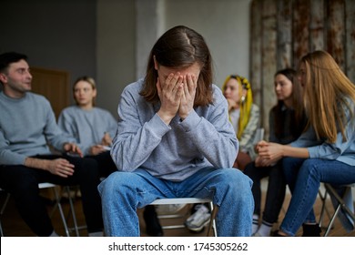 young male suffer from alcohol addiction, cry, sit apart from group of people gathered in the club of anonymous alcoholics