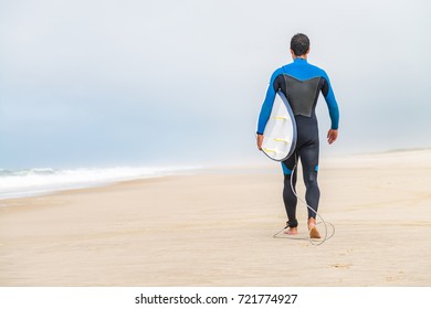 Young male surfer wearing wetsuit, holding surfboard under his arm, walking on beach after morning surfing session. - Powered by Shutterstock