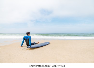 Young male surfer wearing wetsuit, sitting beside his surfboard on beach after morning surfing session. - Powered by Shutterstock