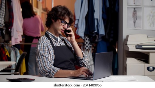 Young Male Tailor Discuss Order With Client On Cellphone At Workshop. Portrait Of Online Store Manager Having Phone Call With Customer Sitting At Desk In Warehouse