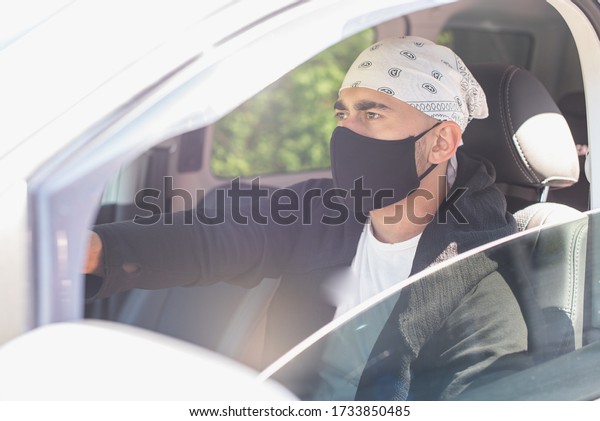 A young\
male taxi driver in a black protective medical mask with a bandana\
and a dark jacket behind the wheel looks straight ahead. Quarantine\
concept. Virus. Pandemic.\
Coronavirus.