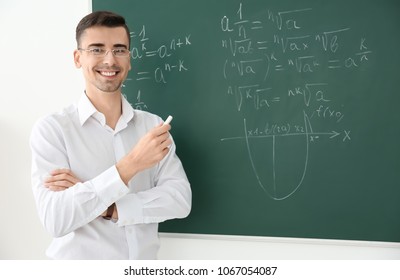 Young Male Teacher With Chalk Near Blackboard In Classroom