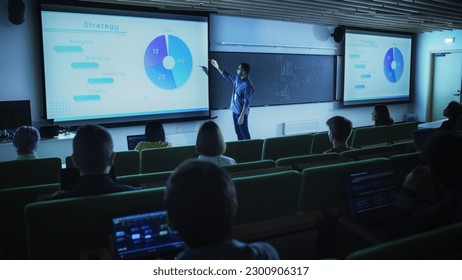 Young Male Teacher Giving a Business Strategy Lecture to Diverse Multiethnic Group of Female and Male Students in Dark College Room. Projecting Slideshow with Comparison Charts and Commercial Data - Powered by Shutterstock