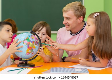 Young Male Teacher Showing His Little Sutdents A Globe, Teaching Geography At Elementary School. Group Of Little Kids Enjoying Learning Geography