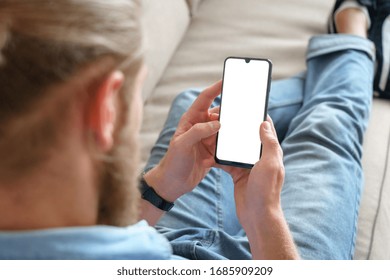 Young Male Tech User Relaxing On Sofa Holding Smartphone Mock Up Blank White Screen. Man Customer Using Cell Phone Mobile Social Media Or Shopping App, Reading News, Sport Betting. Over Shoulder View