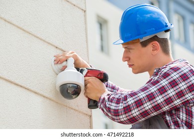Young Male Technician Installing Camera On Wall Using Electric Cordless Drill